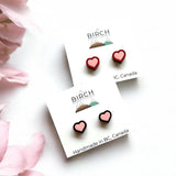 The image is a flatly style image of two pairs of earrings on earring cards. The top card has a pair of wooden heart earrings hand painted pink with a red border. The lower card has a pair of wooden heart studs painted pink with a black border. There is a Birch Street Studio logo on each card and the words at the bottom read, Handmade in BC, Canada.