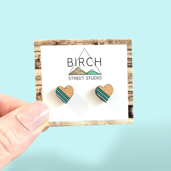 A pair of wooden heart shaped stud earrings. The earrings are hand painted featuring mint green and silver stripes on the left side of the hearts.