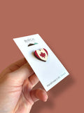 A person's hand holding a white card with a heart-shaped pin featuring a red maple leaf on it.