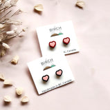 The image is a flatly style image of two pairs of earrings on earring cards. The top card has a pair of wooden heart earrings hand painted pink with a red border. The lower card has a pair of wooden heart studs painted pink with a black border. There is a Birch Street Studio logo on each card and the words at the bottom read, Handmade in BC, Canada.