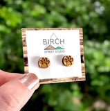 a hand holding a pair of wooden earrings on a card. The wooden earrings are laser cut and engraved with the image of a paw print. The paw print could be either a cat paw or a dog paw. The earrings are hypoallergenic, with surgical steel posts.