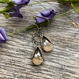 A pair of wooden earrings with mountain designs, resting on a wooden surface with purple flowers in the background.