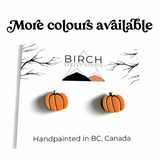 A pair of handpainted orange pumpkin stud earrings displayed on a white Birch Street Studio card, with text reading 'More colours available' and 'Handpainted in BC, Canada'.