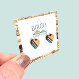 A hand holding a pair of wood heart earrings with rainbow stripes. The rainbow stripes are on half of the earrings, and the colours are red, yellow, green and blue.