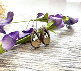 A pair of wooden earrings with mountain designs rests on a wooden surface, surrounded by purple flowers.