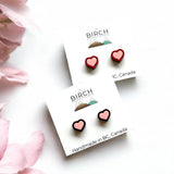 The image is a flatly style image of two pairs of earrings on earring cards. The top card has a pair of wooden heart earrings hand painted pink with a red border. The lower card has a pair of wooden heart studs painted pink with a black border. There is a Birch Street Studio logo on each card and the words at the bottom read, Handmade in BC, Canada.