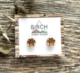 A pair of wood stud earrings with a laser engraved image of paws etched into them. The paws could either be cat paws or dog paws. The earrings are on a card with the Birch Street Studio Logo on them.